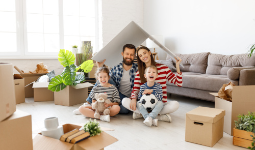 Familie sitzt beim Umzug im hellen Wohnzimmer zwischen Kartons und spielt mit Kindern im Papphaus