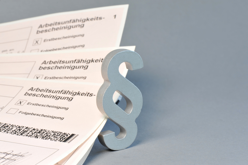 Eine grau-weiße Paragrafen-Zeichenfigur steht neben mehreren Arbeitsunfähigkeitsbescheinigungen.